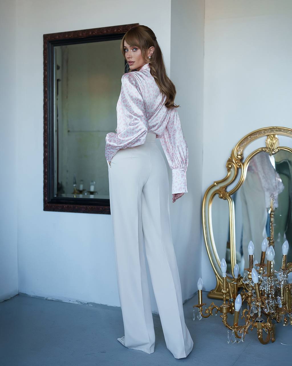 Woman in a white outfit standing in a room with decorative mirrors and a chandelier.
