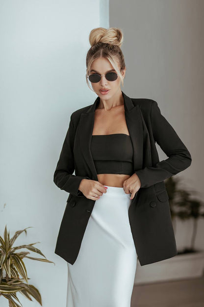 Woman wearing a black blazer over a black top and white pants, standing against a light-colored wall.