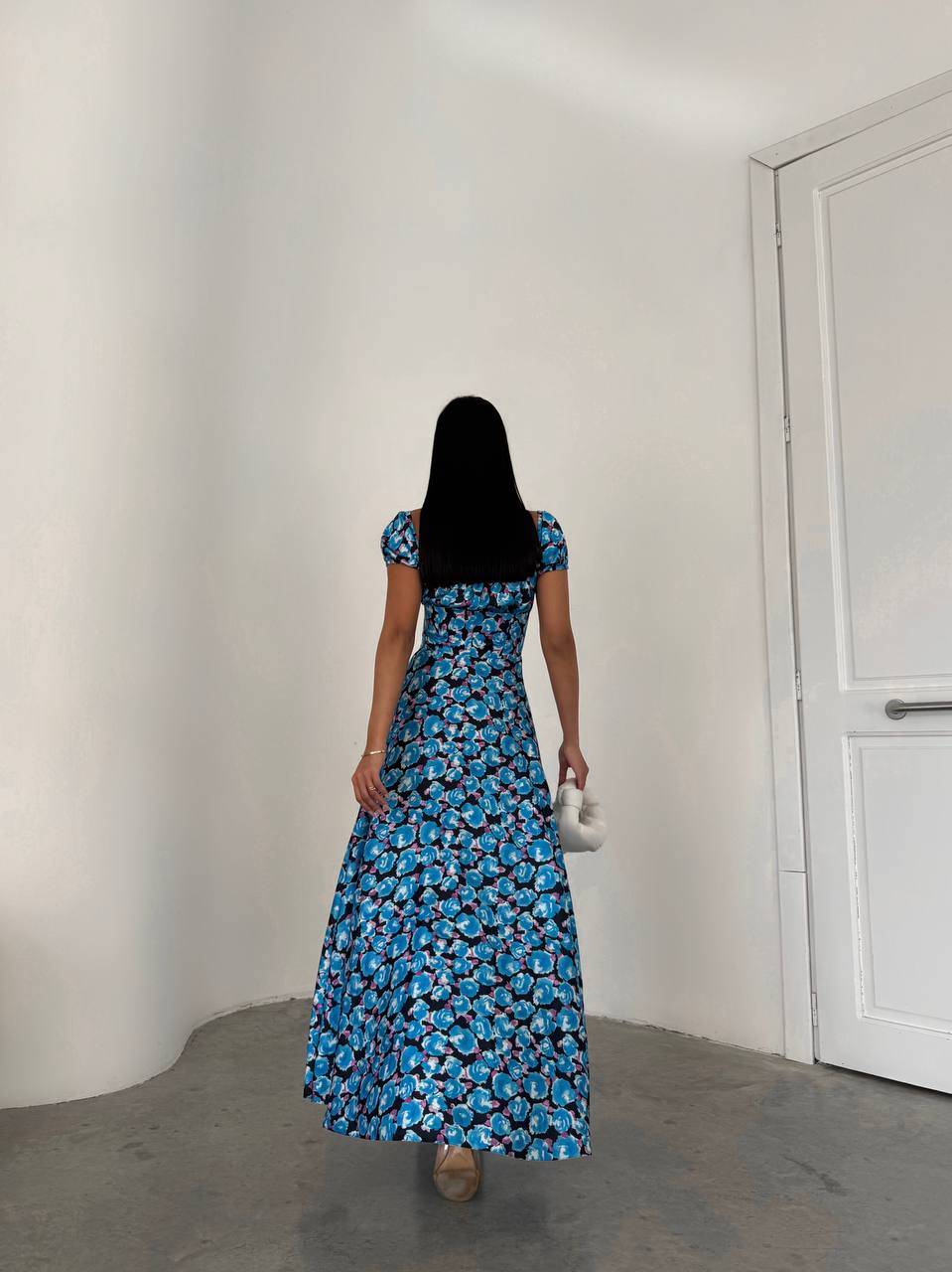 Woman in a blue floral dress standing in a minimalistic room with a white wall and door.