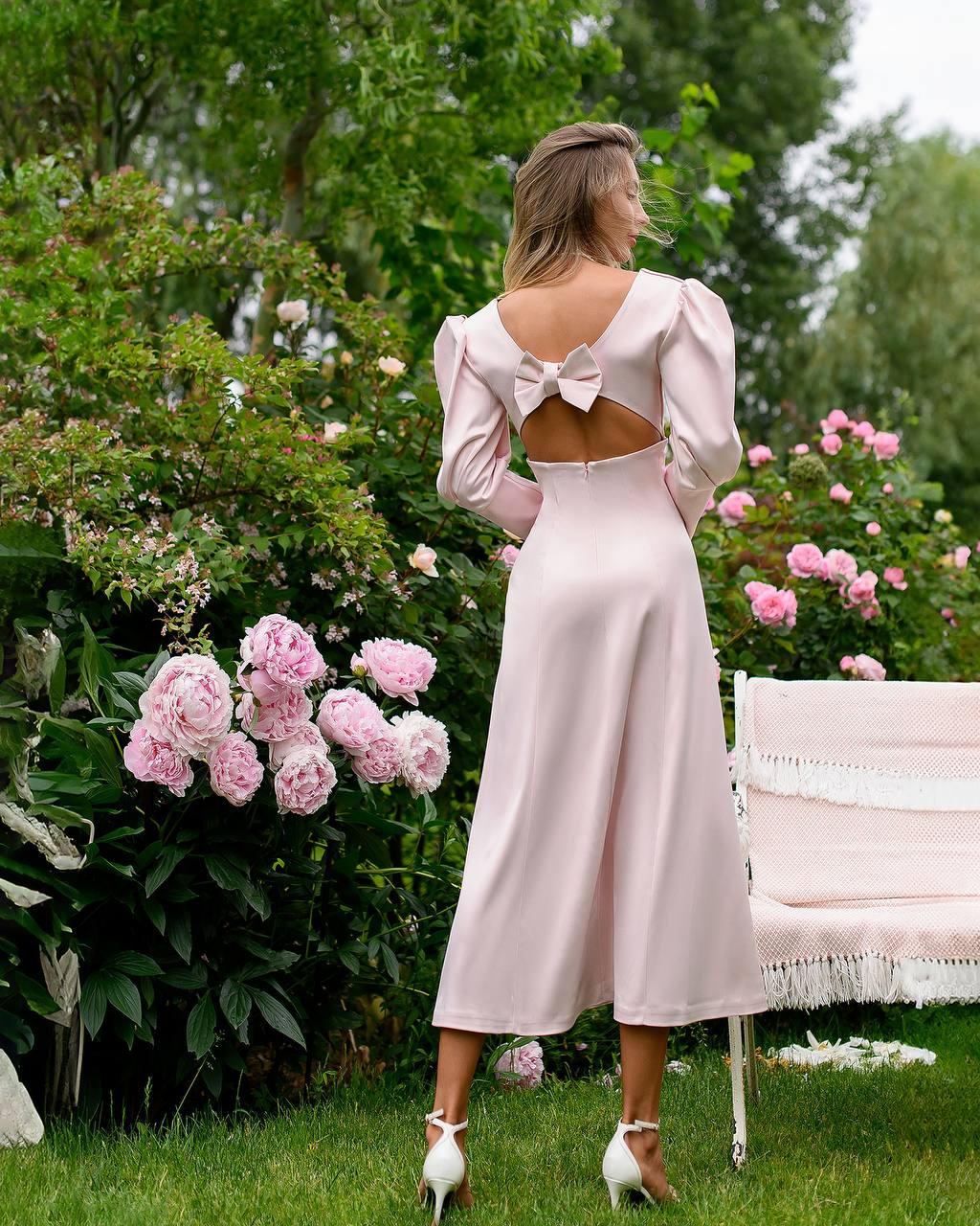 Woman in a light pink dress with cut-out back standing in a garden with pink flowers and a white bench.