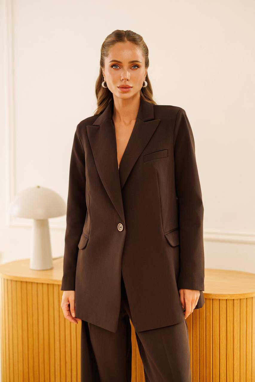 A woman modeling a dark brown tailored blazer with matching trousers, standing in a bright indoor setting with a light wood table and a white lamp in the background.