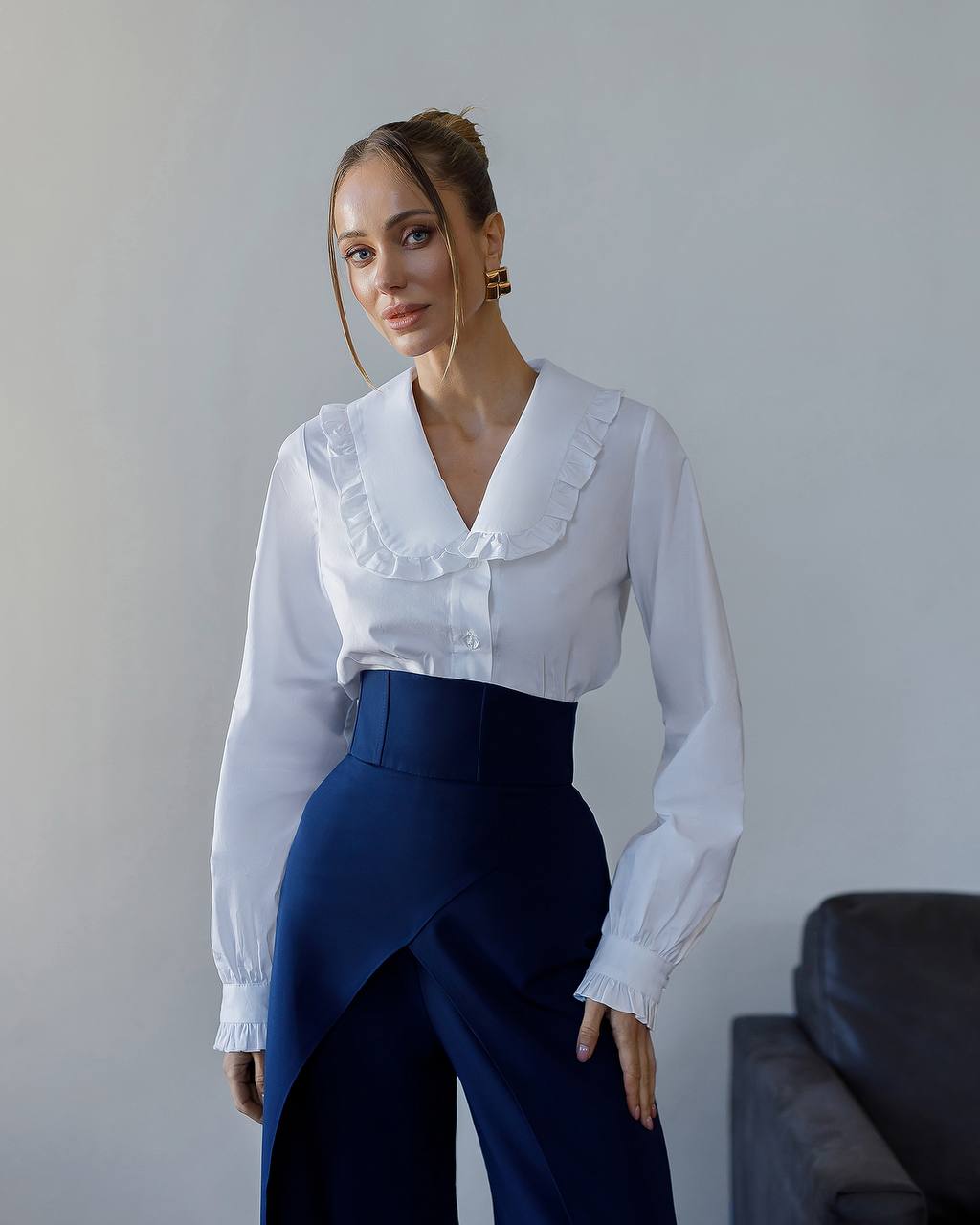 Woman wearing a white blouse with ruffled details and dark blue pants against a plain background