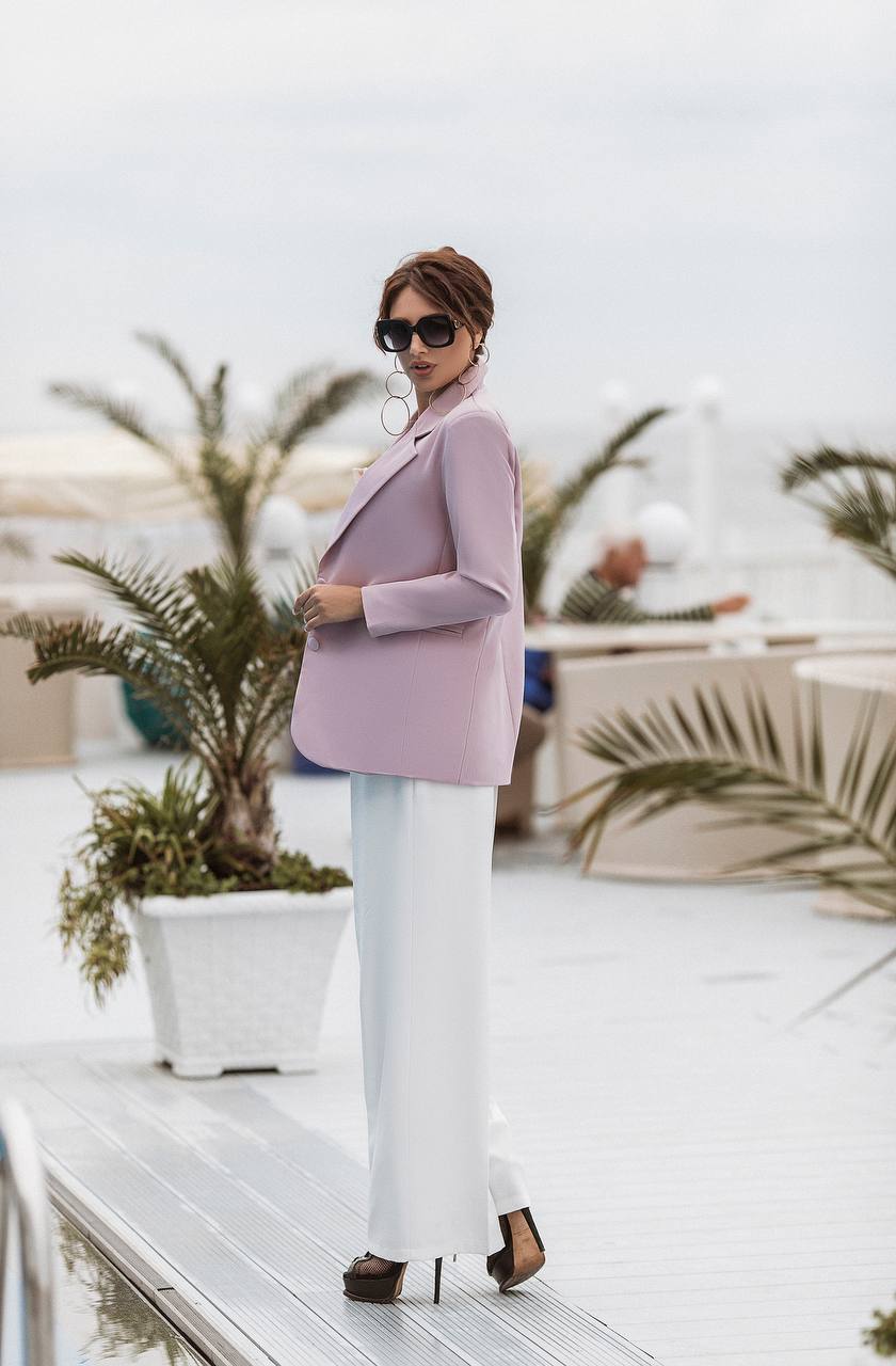 A woman stands on a white deck wearing a lavender blazer, white wide-leg pants, and black heels, with sunglasses and hoop earrings, in front of a blurred outdoor patio background with potted palm trees.