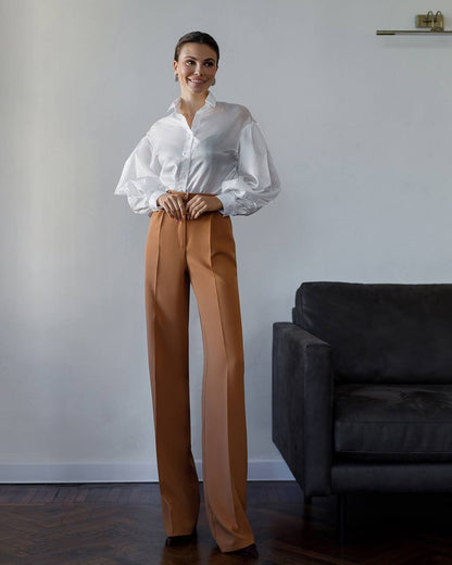 Woman wearing a white blouse and brown pants standing in a room with a gray wall and dark sofa.