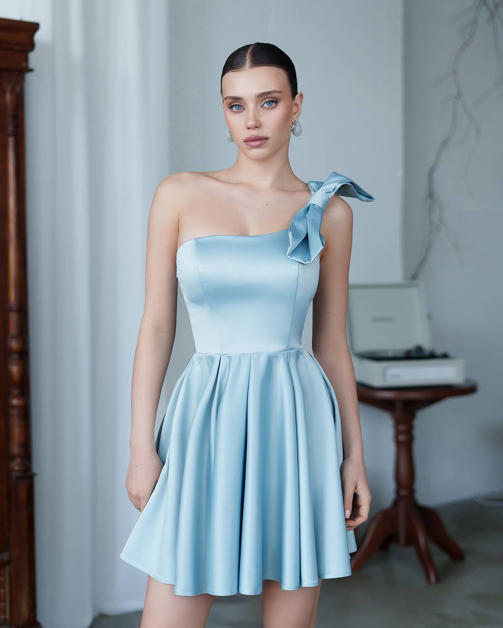 Woman wearing a light blue one-shoulder dress with a bow detail in an indoor setting.