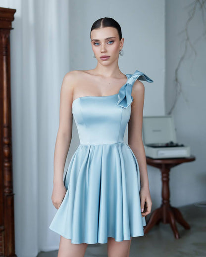 Woman wearing a light blue one-shoulder dress with a bow detail in an indoor setting.