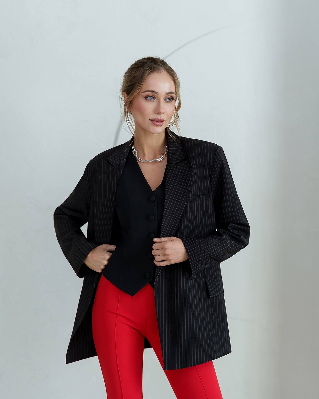 Woman wearing a black pinstripe jacket over a black top with red pants against a light gray background
