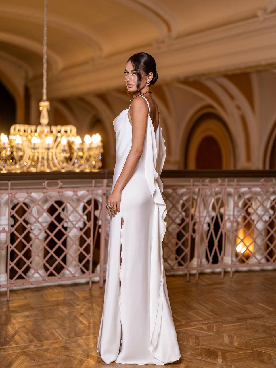 Woman in a white dress standing in an elegant indoor setting with chandeliers.
