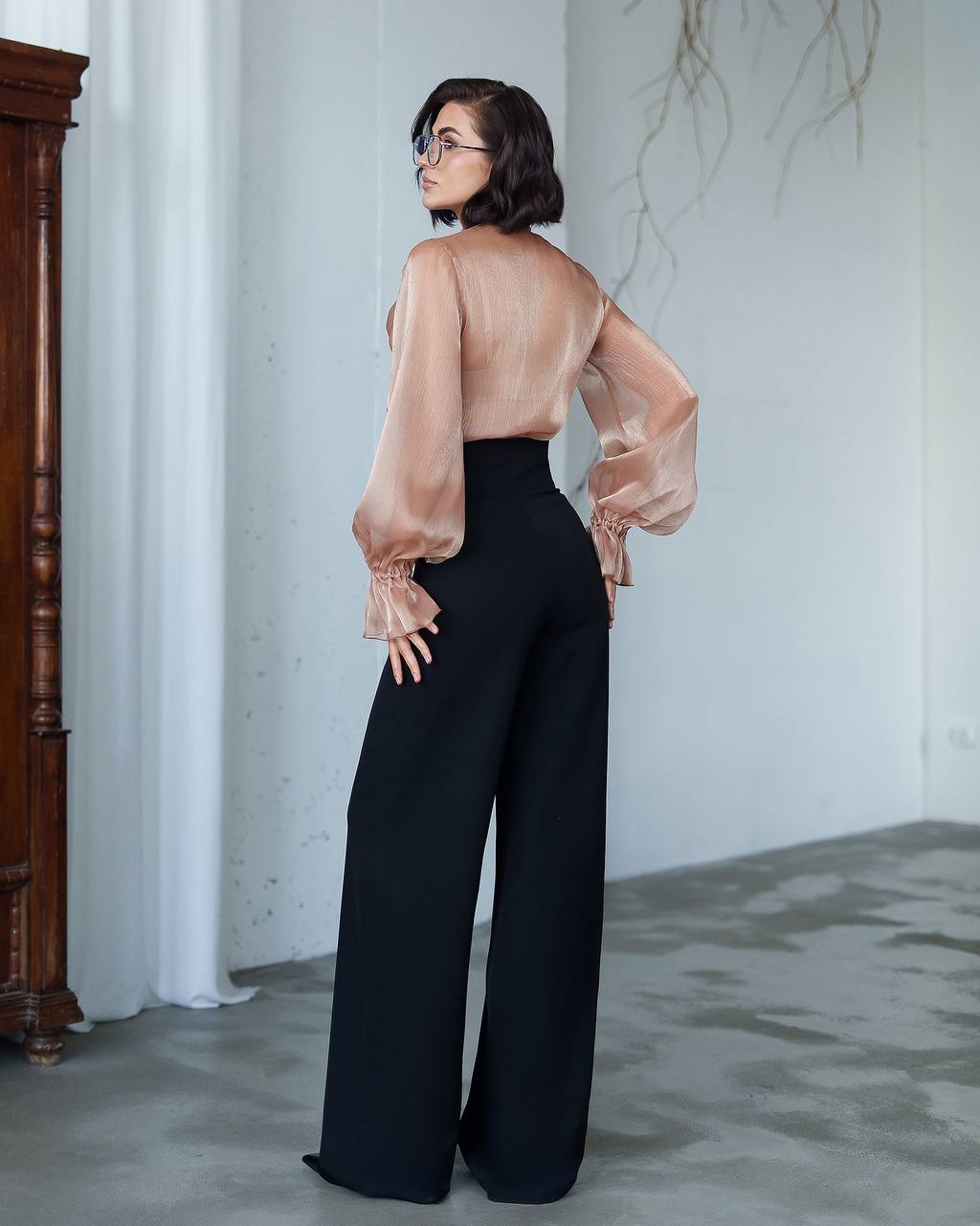 Woman wearing a sheer beige blouse and black pants in a minimalistic room.