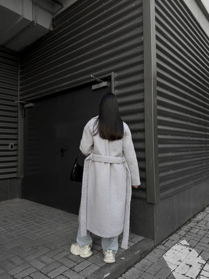 Person in a long coat standing in front of a dark door with horizontal slats.