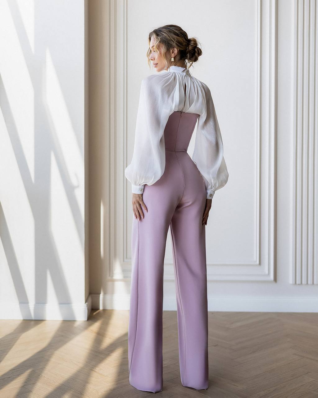 Woman wearing a white blouse and lavender pants in a room with sunlight filtering through.