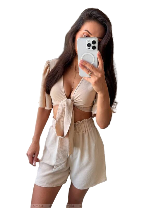 Woman taking a mirror selfie wearing a beige top and white shorts on a white background