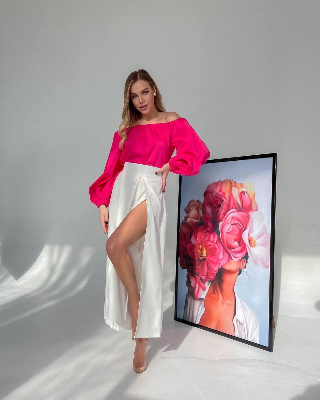 Woman in a bright pink top and white skirt standing next to a framed artwork on a gray background