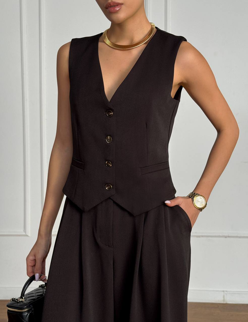 Woman wearing a black sleeveless vest and skirt set against a white wall.