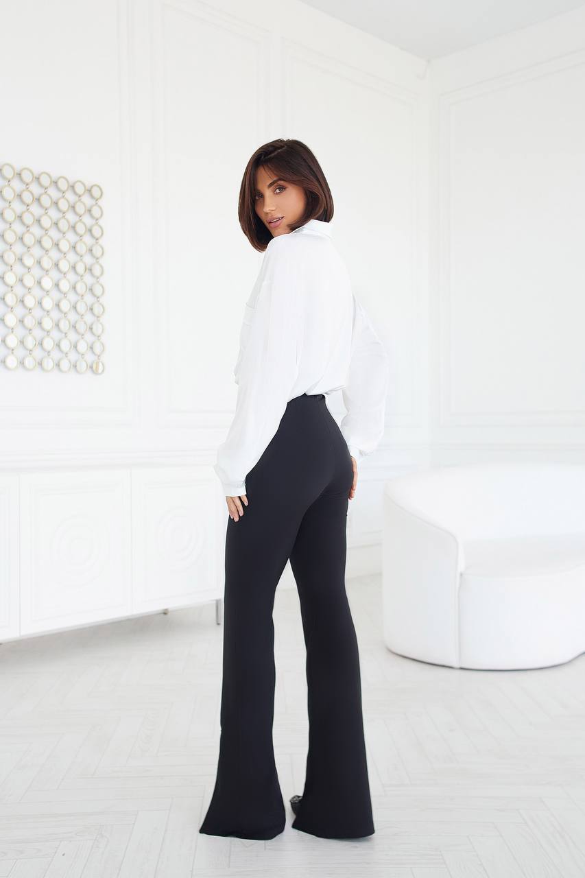 Woman wearing a white blouse and black pants in a minimalistic room.
