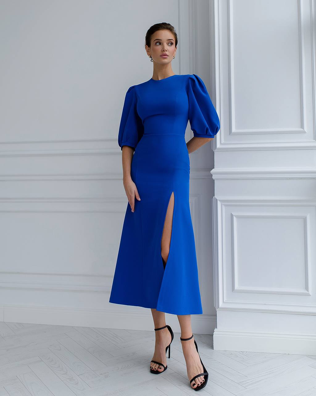 Woman wearing a blue dress with puff sleeves against a white wall.