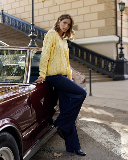 Woman in a yellow sweater and blue pants leaning against a vintage car on a city street.