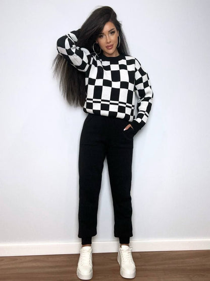 Person wearing a black and white checkered sweater with black pants and white shoes against a white wall.