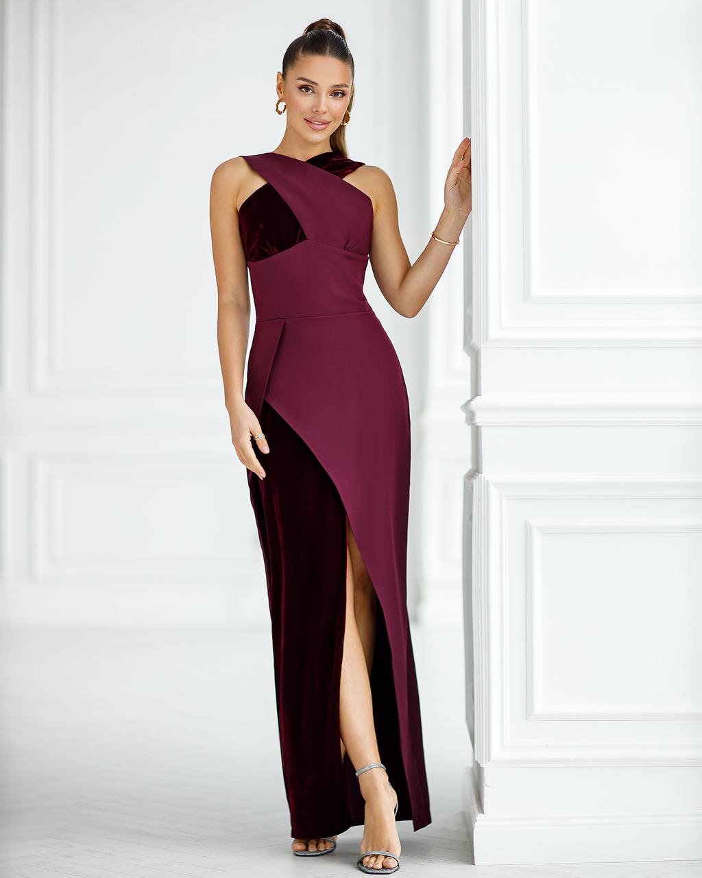 A woman models a floor-length evening gown in a bright white paneled studio setting. The dress is deep burgundy with a velvet-like textured bodice panel and a high thigh slit.