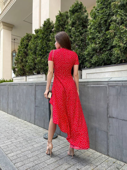 Woman in a red polka dot dress walking outdoors near a building with greenery.
