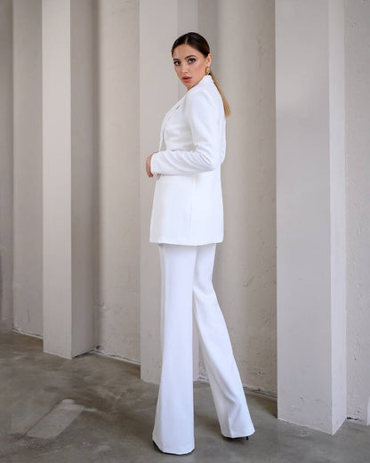Woman wearing a white suit standing against a plain wall.