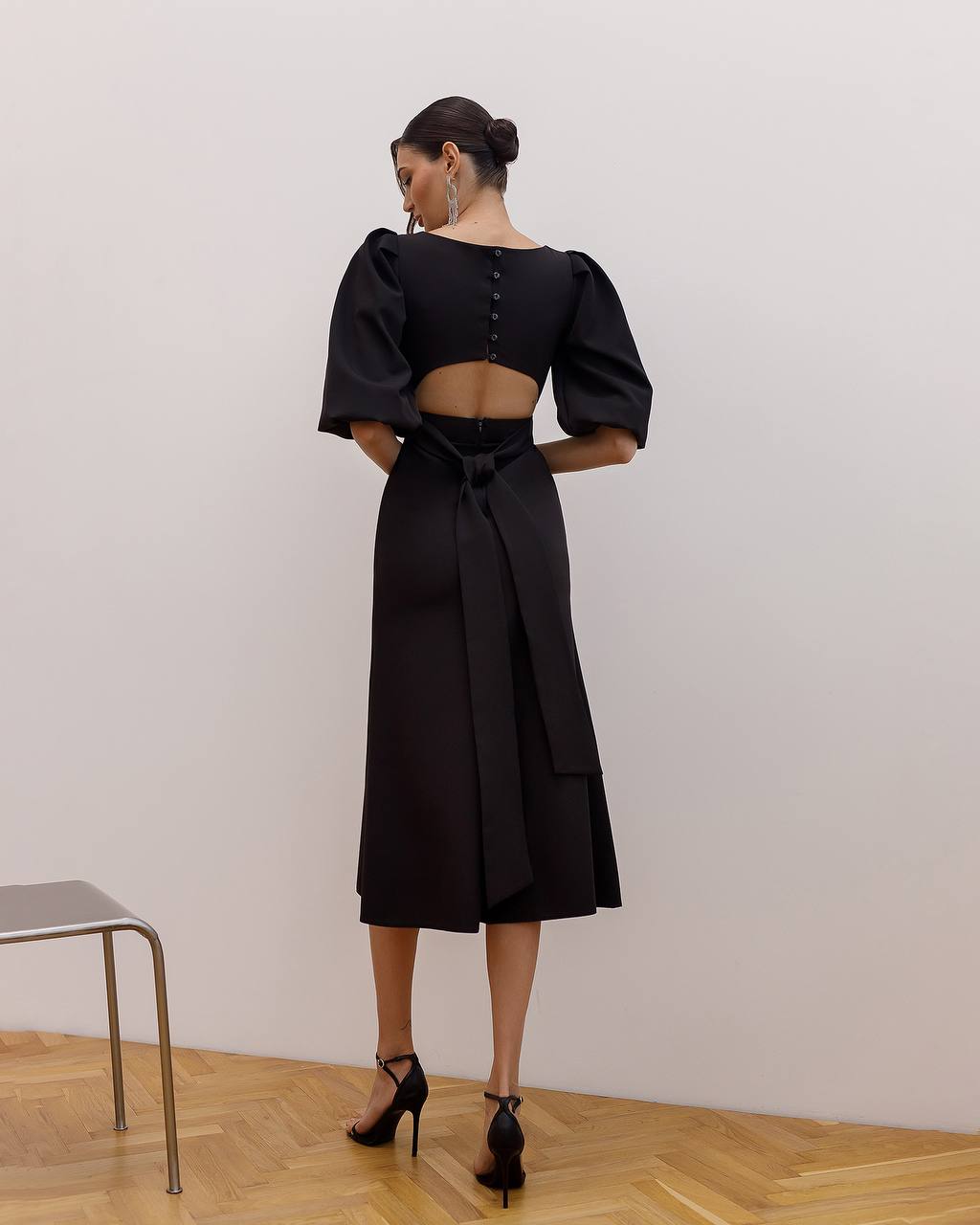 Woman wearing a black outfit with puffed sleeves and a belt, standing against a plain wall.