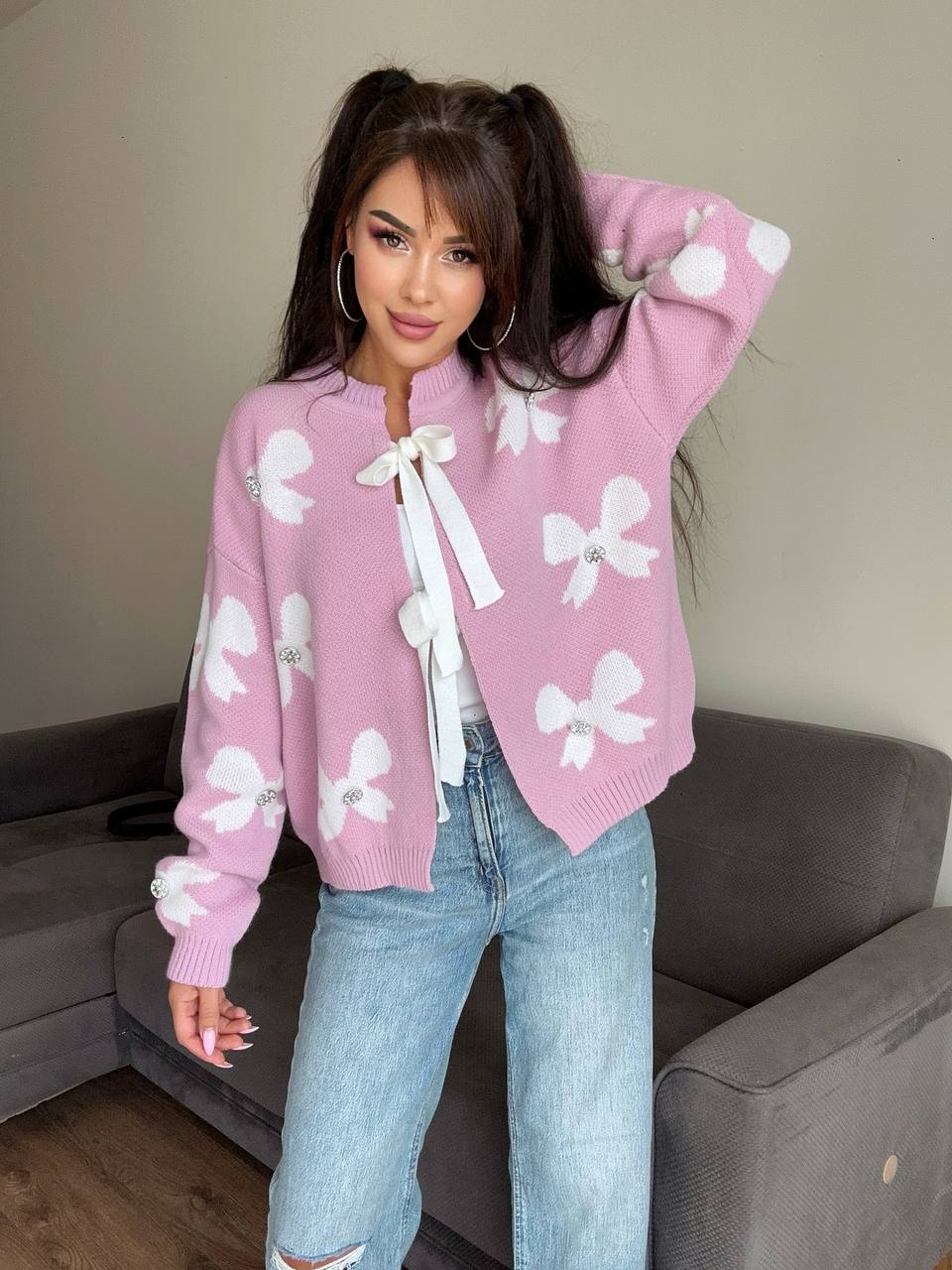 Woman wearing a pink sweater with white bow patterns and light blue jeans indoors.