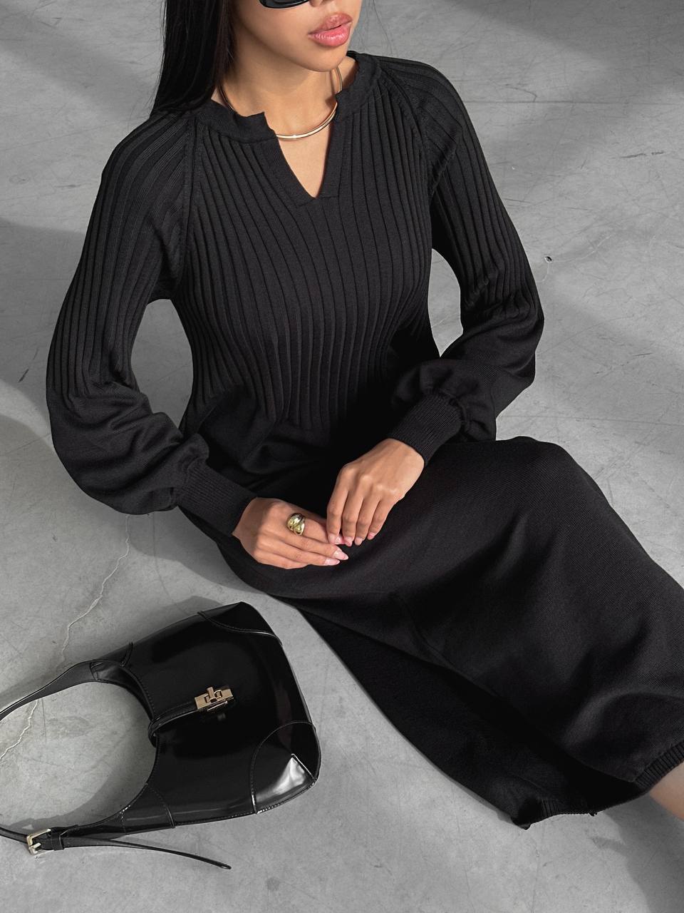 Person wearing a black ribbed dress sitting on a gray surface with a black handbag beside them.