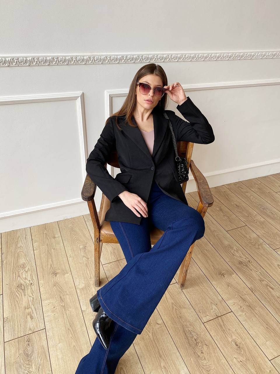 Woman sitting on a chair wearing a black blazer and blue jeans in an elegant room.