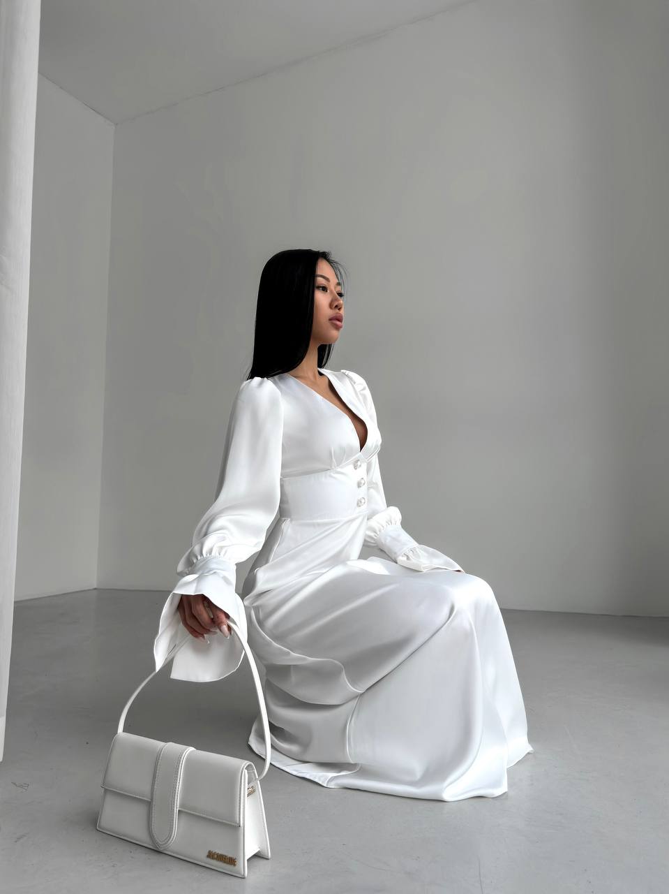Woman in a white dress sitting on the floor with a matching handbag in a minimalistic setting.