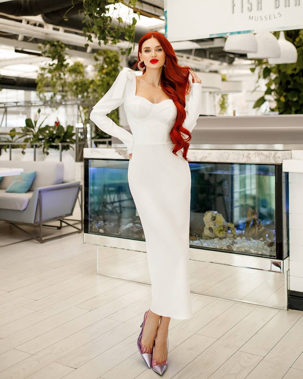 Woman in a white dress posing in an indoor setting with plants and furniture.