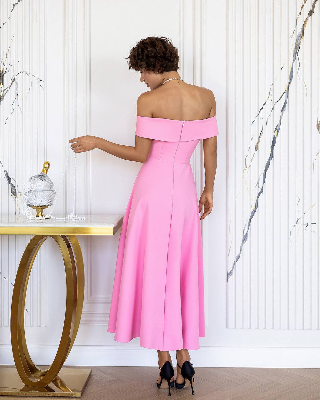 Woman in a pink off-shoulder dress standing in a room with a marble wall and gold table.