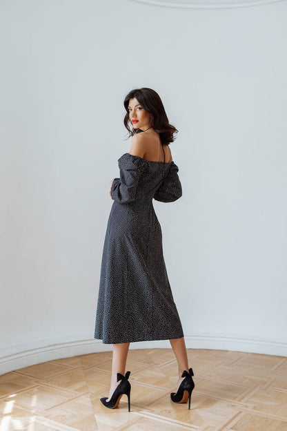 Woman in a black polka dot dress standing in a room with wooden flooring and white walls.