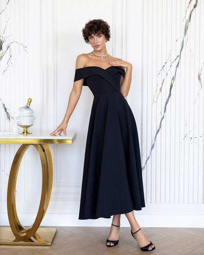 Woman in a black off-shoulder dress standing next to a gold table against a white marble wall.