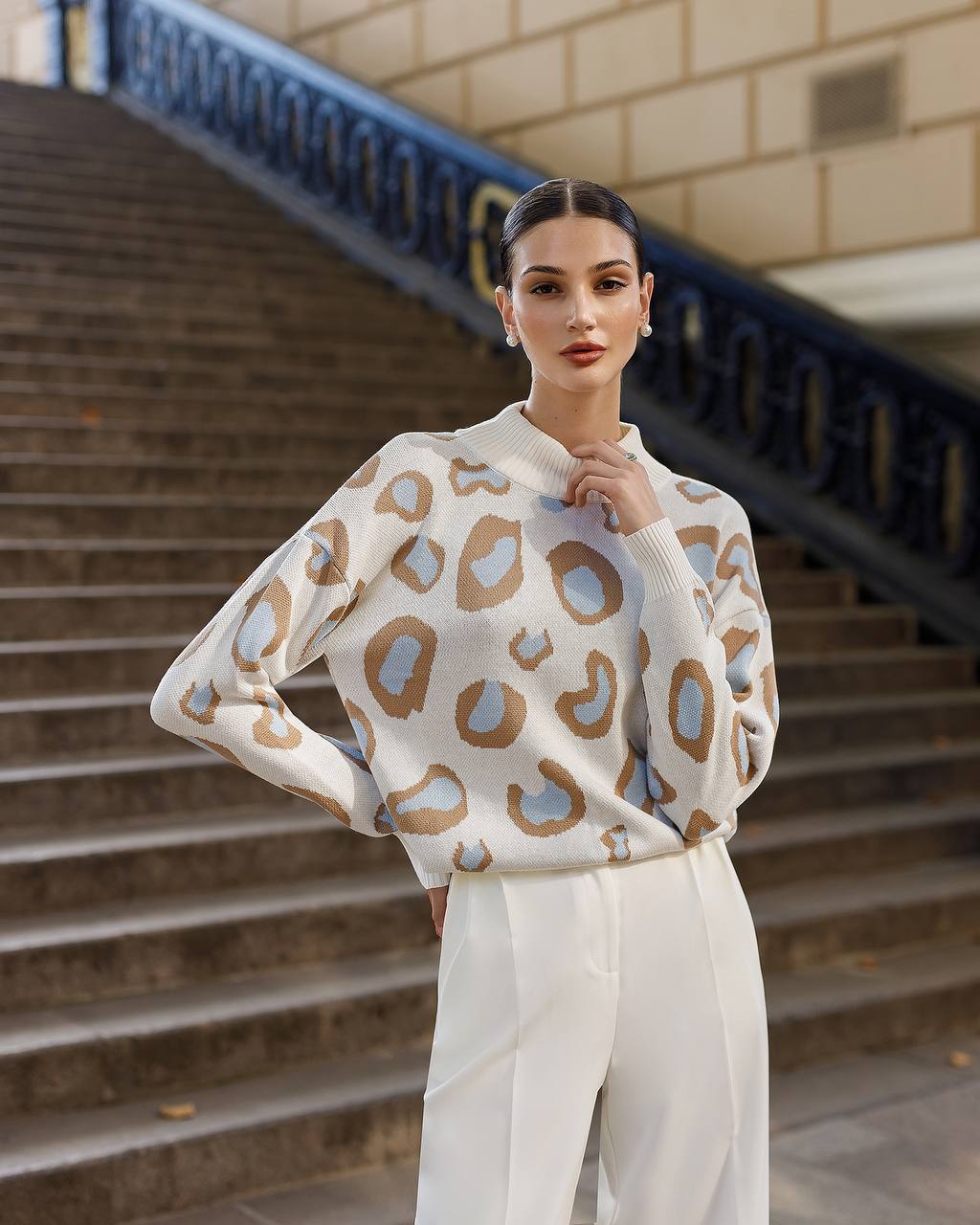 Woman wearing a patterned sweater and white pants standing on steps.