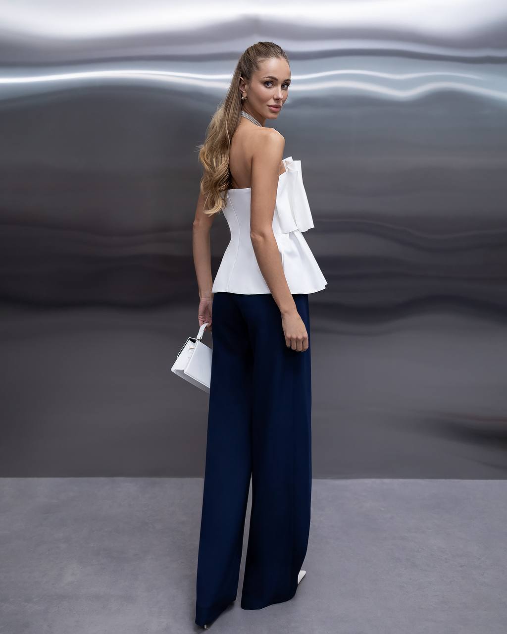 A woman stands with her back to the camera wearing a white strapless ruffled top and navy wide-leg trousers, holding a small white handbag against a dark gray abstract studio backdrop.