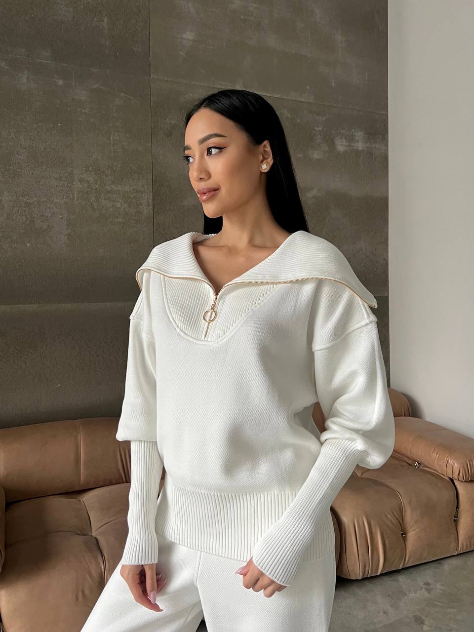Woman wearing a white sweater with a gold zipper in a room with a brown couch.