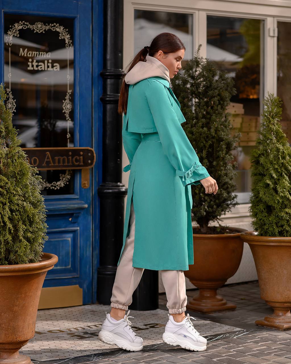 Woman in a teal coat standing outside a building with potted plants.