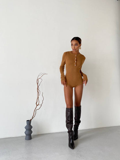 Woman in a brown bodysuit and black knee-high boots standing next to a vase with a plant on a white background