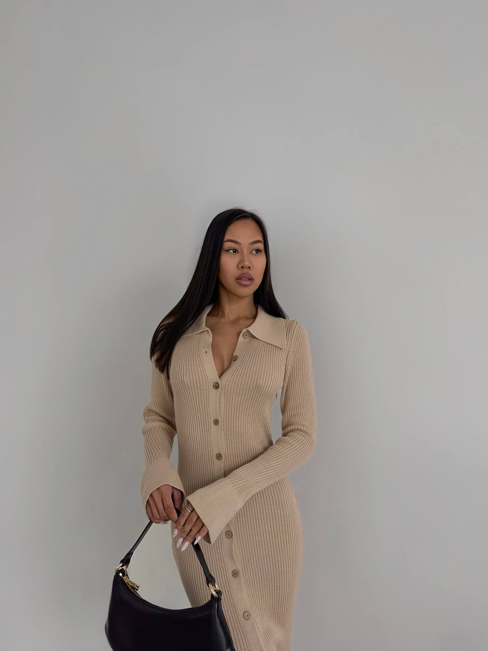 Woman wearing a beige ribbed dress holding a black handbag against a plain background
