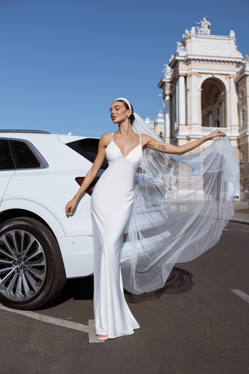 Woman in a white wedding dress standing next to a white car with a classical building in the background
