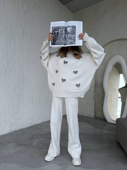 Person wearing a white sweater with heart patterns and white pants, holding a book in front of their face.