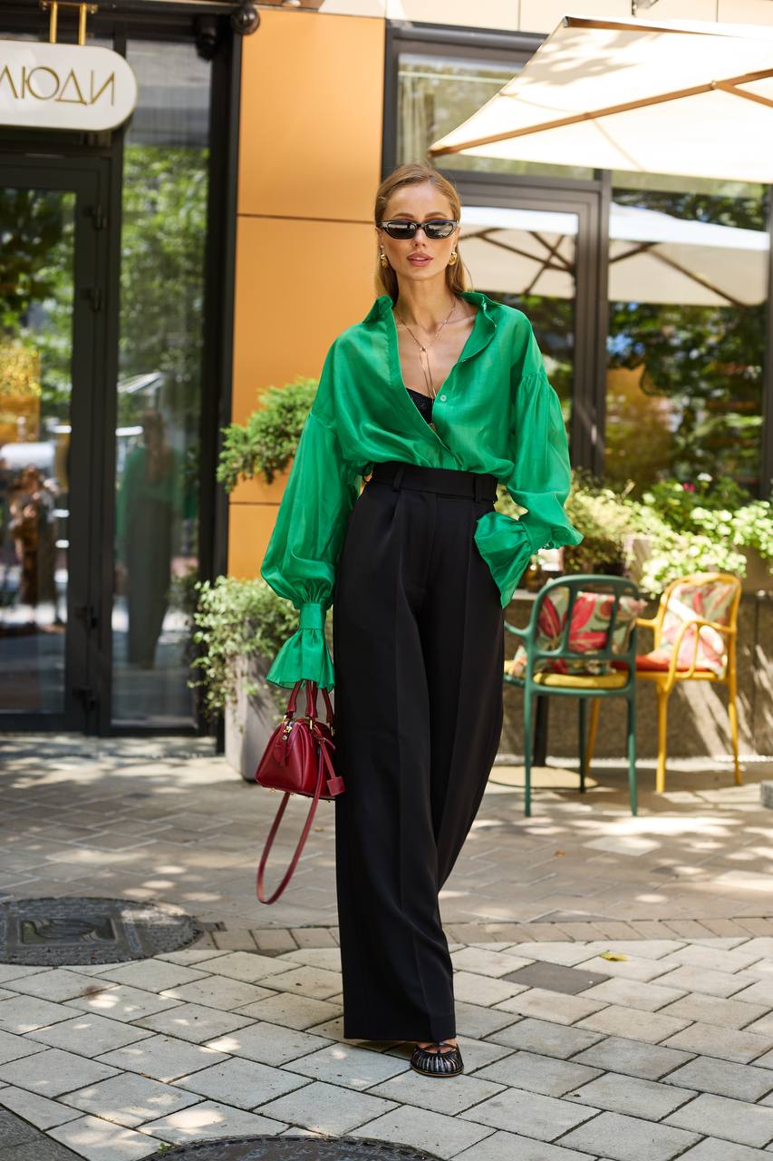 Woman in a green blouse and black pants standing on a street with a building and chairs in the background.
