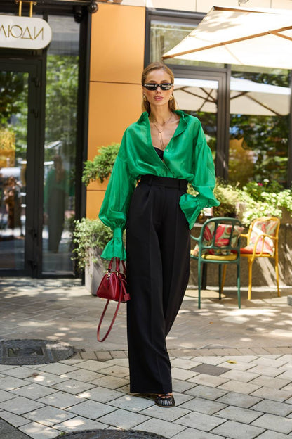 Woman in a green blouse and black pants standing on a street with a building and chairs in the background.