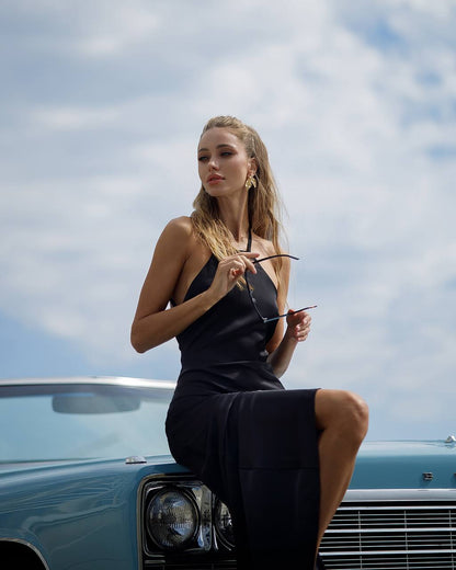 Woman in a black dress sitting on a classic car with a blue sky background