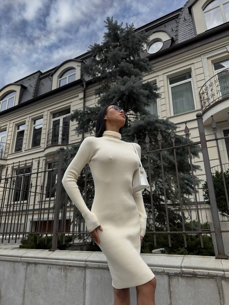 Woman in a white dress standing in front of a European-style building with trees and sky.