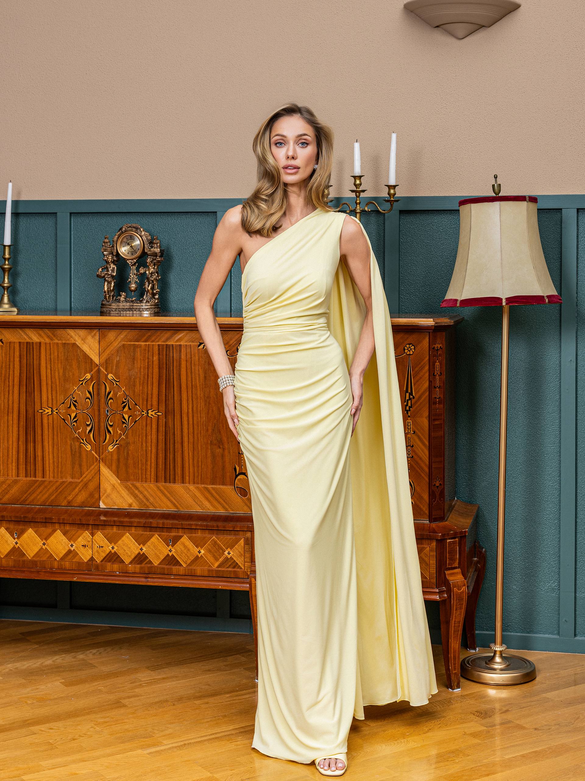 Woman in a light yellow one-shoulder dress standing in an elegant room with wooden furniture and a lamp.