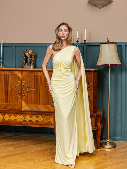 Woman in a light yellow one-shoulder dress standing in an elegant room with wooden furniture and a lamp.