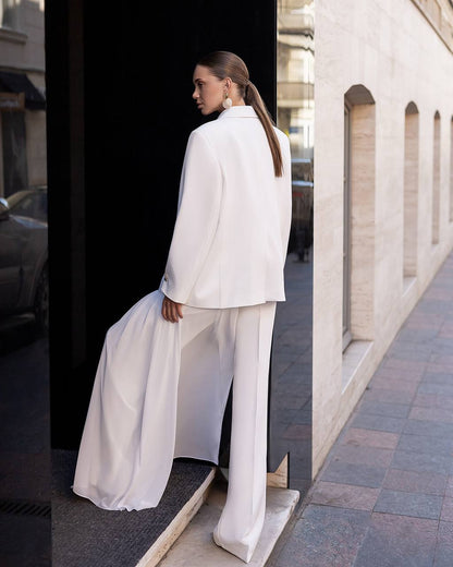 A woman wearing a white satin-like two-piece outfit with a structured blazer and wide-leg trousers, standing on a sidewalk beside a building with arched windows.