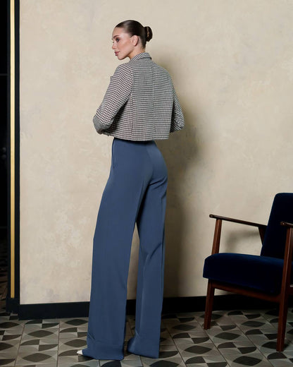 Woman in a checkered jacket and blue pants standing against a beige wall with a chair in the corner.